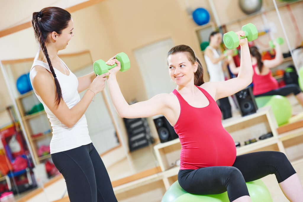 A trainer with a pre and postnatal fitness certification coaching a pregnant client with weights on a birth ball. 